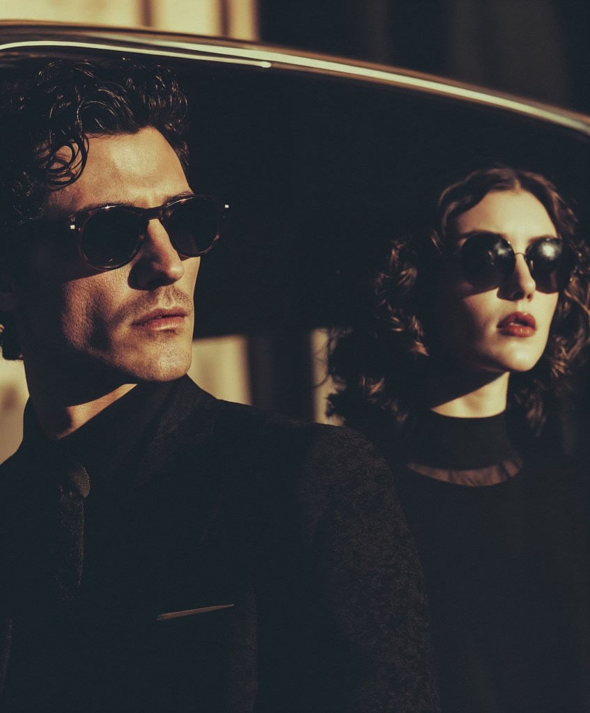 Dos figuras con estilo clásico, gafas de sol y aire cinematográfico emergen entre luces dramáticas y sombras profundas.
